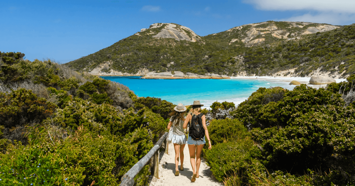 Two women walking on a sandy path through green bushes towards a bright turquoise bay surrounded by hills.