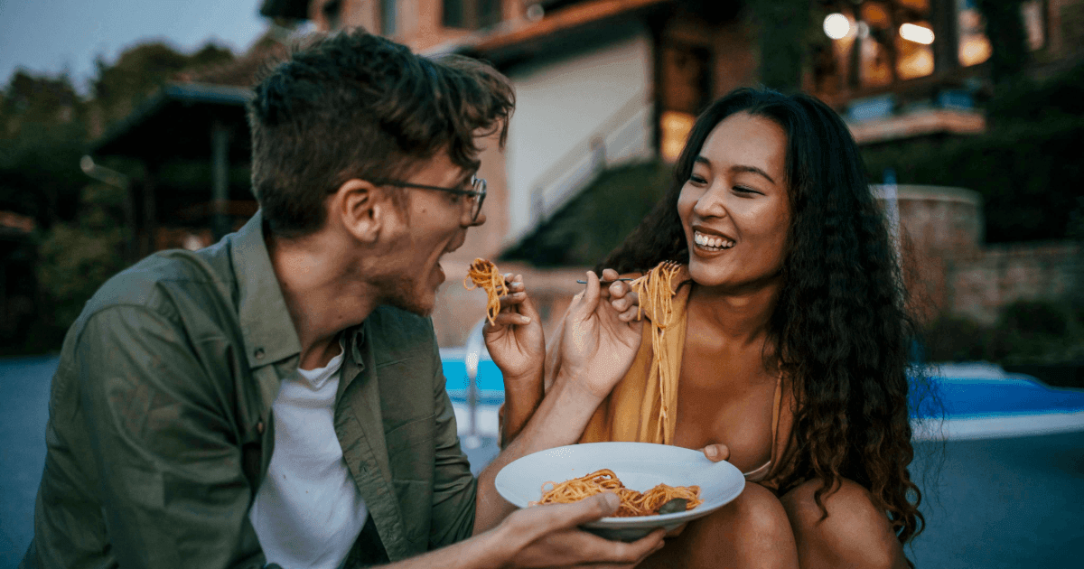 Two people sitting outdoors, sharing a plate of spaghetti, with a blurred and greenery-filled background.